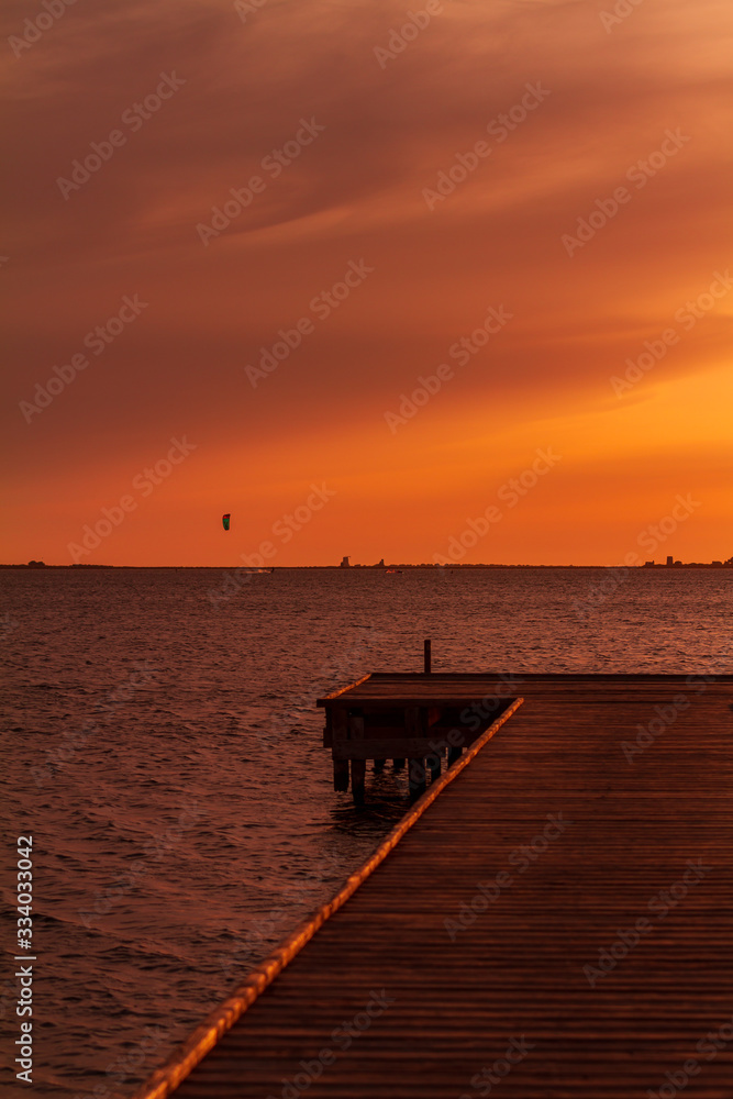 Wooden pier on a warm light during sunset, with orange sky and a Kite surf in the background