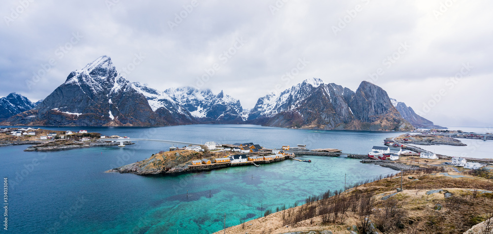 Panorama of the island of sakrisoy and hamnoy in Lofoten islands in Norway. Turquoise water infront of yellow guest houses and snow covered mountains in the background.