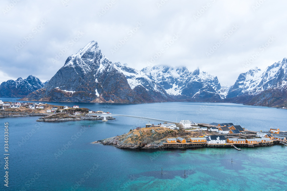 Sakrisoy island in Lofoten, Norway. Turquoise water infront of yellow guest houses with bridge over to Hamnoy island with snow covered mountain Olstind in the background, foggy weather.