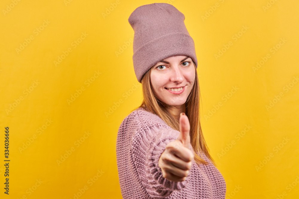 Young beautiful girl showing thumbs up gesture. woman is wearing a warm sweater and hat. isolated on a yellow background. Young beautiful girl showing thumbs up gesture. woman is wearing a warm sweater and hat. isolated on a yellow background.