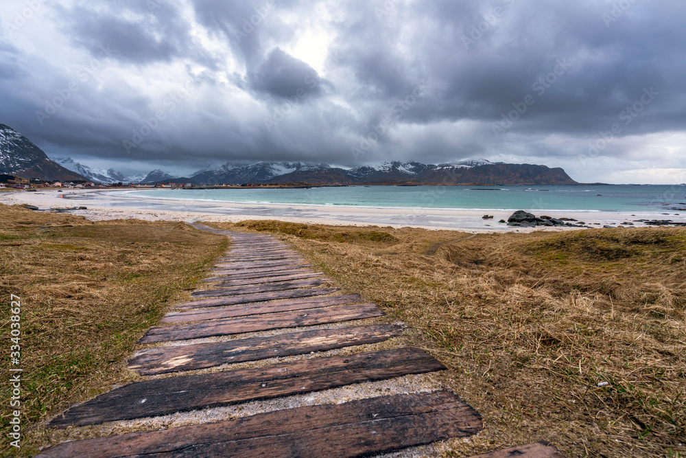 Wooden path towards Ramberg beach in Lofoten during rainy weather and blue hour. Snow covered mountains in the background and the village of Flakstad.