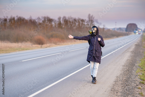 COVID-19. A girl in dark clothes and a gas mask outside the city on a cloudy day stands on the edge of the road.