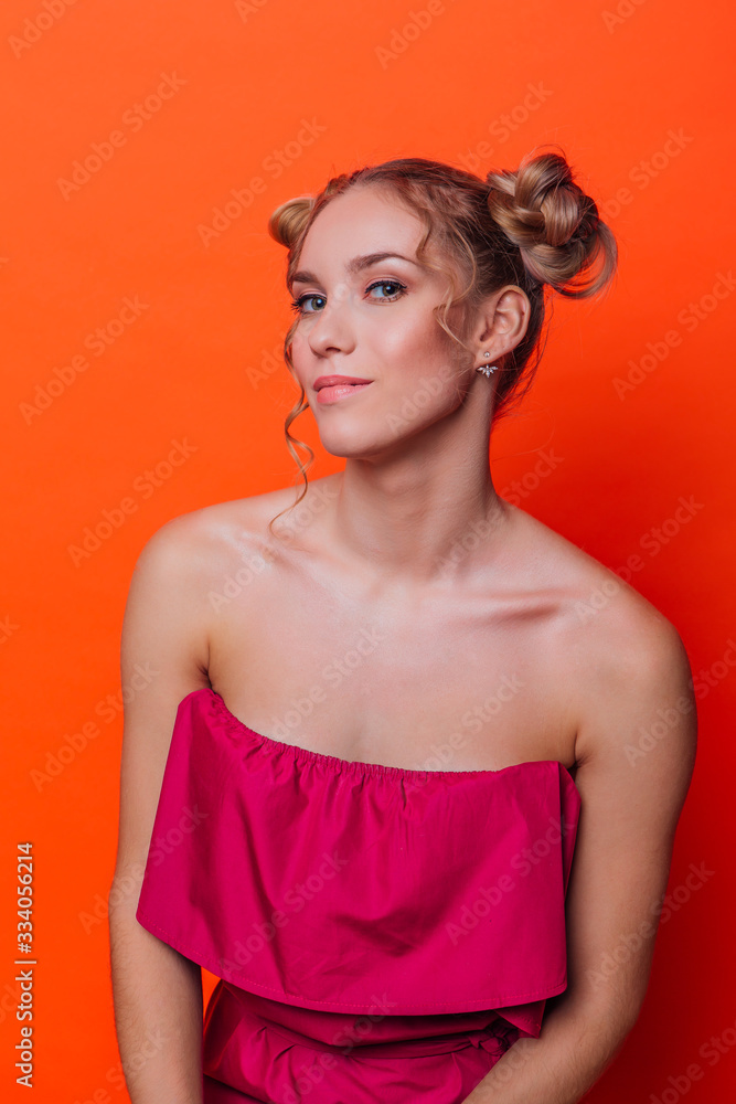 The girl in a red dress on a orange background in the studio. Blonde girl with two hair knots looking to the camera and smiling.