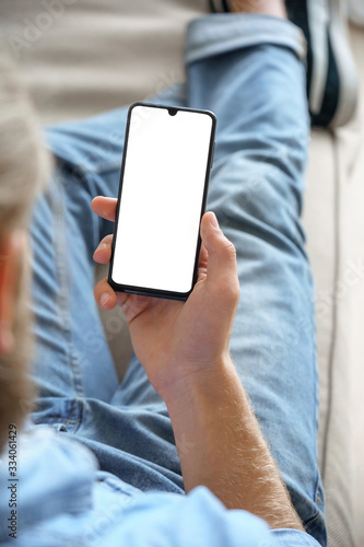 Young man customer choosing buying order in shopping app, reading social media news, texting mobile sms message in chat, using smart phone mock up blank screen sit on sofa. Vertical over shoulder view