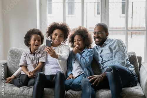 African ethnicity couple with little kids resting on couch having fun holding smart phone using new free cool application  making self-portrait capture moment  enjoy distant talk by video call concept