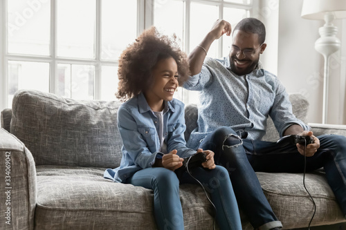 African ethnicity father spend time with little daughter, family sit on couch holding joypads competing in playstation video games, dad loses excited kid girl win. Weekend activity, free time concept