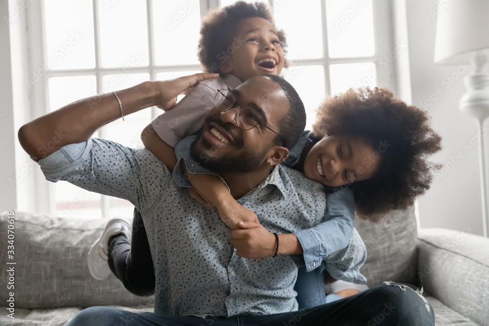 Happy African family at home, cute son and daughter hanging on daddy back, father fool around with little son and daughter piggy back siblings enjoy active time together seated on sofa in living room - obrazy, fototapety, plakaty 