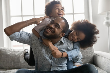 Happy African family at home, cute son and daughter hanging on daddy back, father fool around with little son and daughter piggy back siblings enjoy active time together seated on sofa in living room