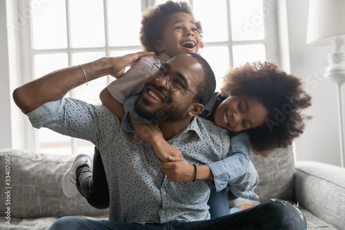 Happy African family at home, cute son and daughter hanging on daddy back, father fool around with little son and daughter piggy back siblings enjoy active time together seated on sofa in living room photo