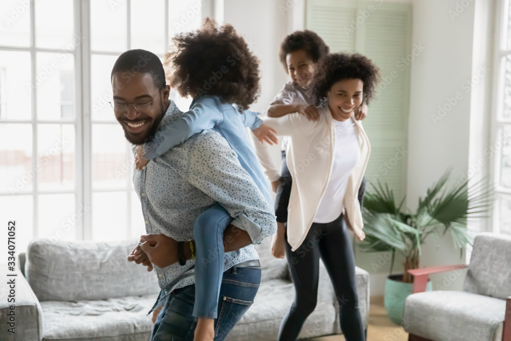 Fototapeta premium Cheerful African couple play with children running piggy back riding little kids celebrating moving day at new first modern home. Family have fun enjoy free time together leisure active games concept