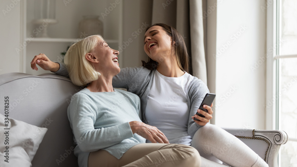 Overjoyed mature mother and millennial grown-up daughter relax on sofa in living room laugh watch funny video on smartphone, happy senior mom and adult girl child have fun using cellphone together
