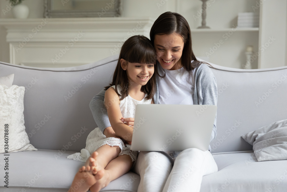Happy loving young mother sit relax on sofa with little daughter watch video on laptop together, smiling millennial mom or nanny rest on couch in living room with small girl child using computer Happy loving young mother sit relax on sofa with little daughter watch video on laptop together, smiling millennial mom or nanny rest on couch in living room with small girl child using computer