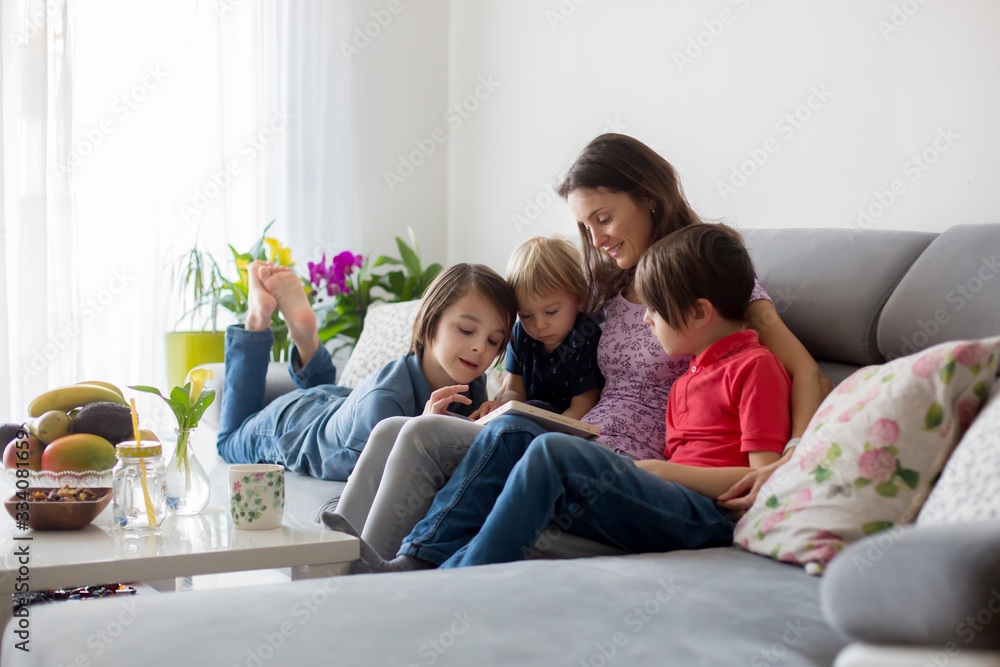 Young woman, mother with three kids, reading a book at home, hugging and laughing Young woman, mother with three kids, reading a book at home, hugging and laughing