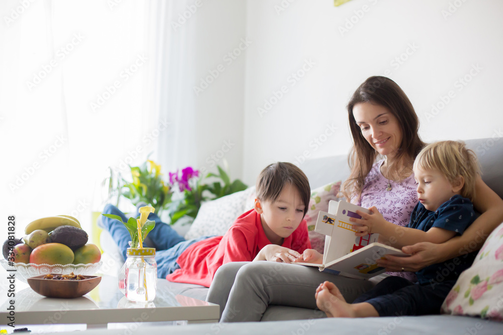 Young woman, mother with three kids, reading a book at home, hugging and laughing Young woman, mother with three kids, reading a book at home, hugging and laughing