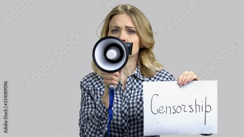 Portrait of sad woman with taped mouth tries to speak in megaphone. Holding babber with censorship word. Grey isolated background. photo