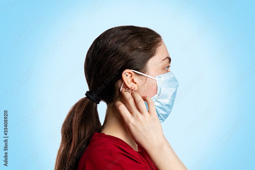 Portrait of a brunette woman in a medical mask and a red t-shirt, on a light blue background. Copy space. View at profile. Concept of pandemic and protection against viral infection