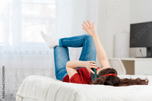 A young woman is lying on the bed on her back and chatting on the phone waving her hand. White interior of the room in the background. Concept of communication and social networks photo