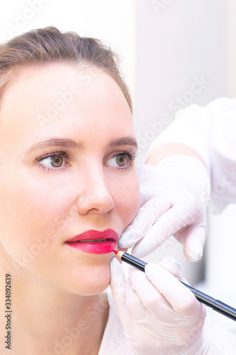 Young woman having permanent makeup on her lips at the beauticians salon. Permanent Makeup (Tattoo). drawing a contour with a white lip pencil. vertical photo