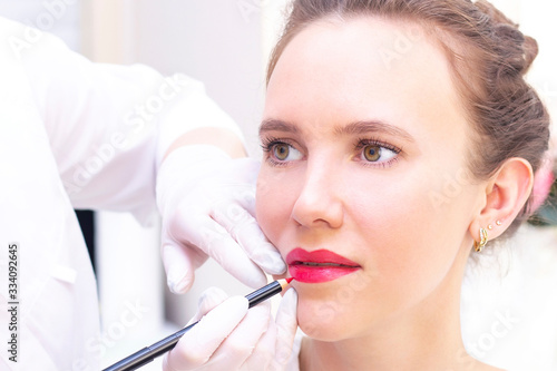 Young woman having permanent makeup on her lips at the beauticians salon. Permanent Makeup (Tattoo). drawing a contour with a white lip pencil