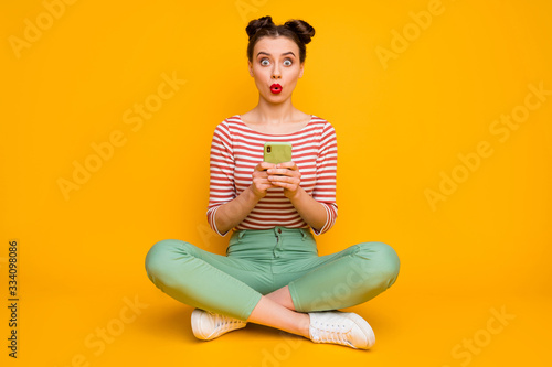 Full length photo of attractive lady speechless sit floor legs crossed hold telephone shocked wear striped red white shirt green pants shoes isolated yellow color background