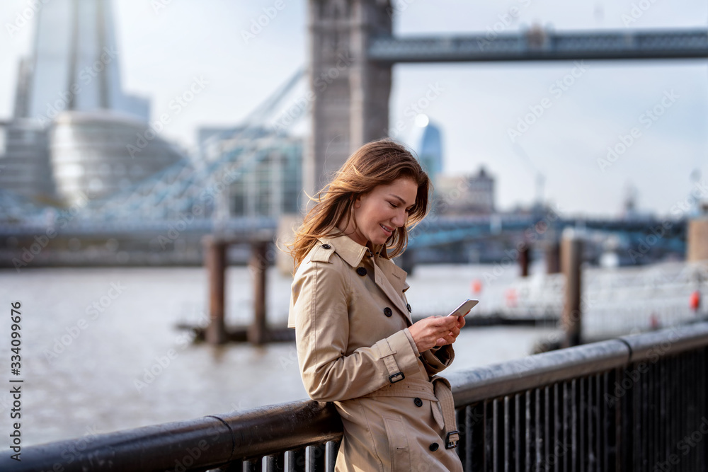 Eine attraktive Frau im Trench Coat Mantel steht an der Themse vor der Skyline von London und liest Emails auf ihrem Mobiltelefon