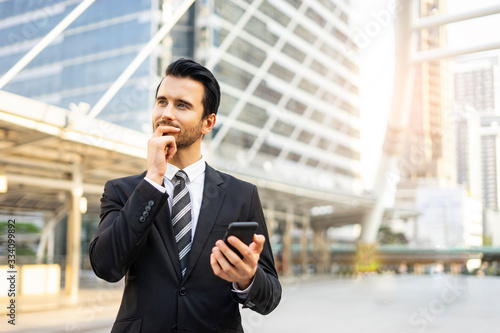 Close up of a caucasian businessman with his hand on the chin thinking, using a smartphone surfing on internet, handsome looking and wearing a suit and tie within an urban city structure in background