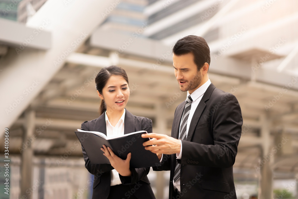 asian businesswoman holding a note book with caucasian businessman turning on the page and reading about business work, outdoor in the city district with urban architectural structure in background asian businesswoman holding a note book with caucasian businessman turning on the page and reading about business work, outdoor in the city district with urban architectural structure in background
