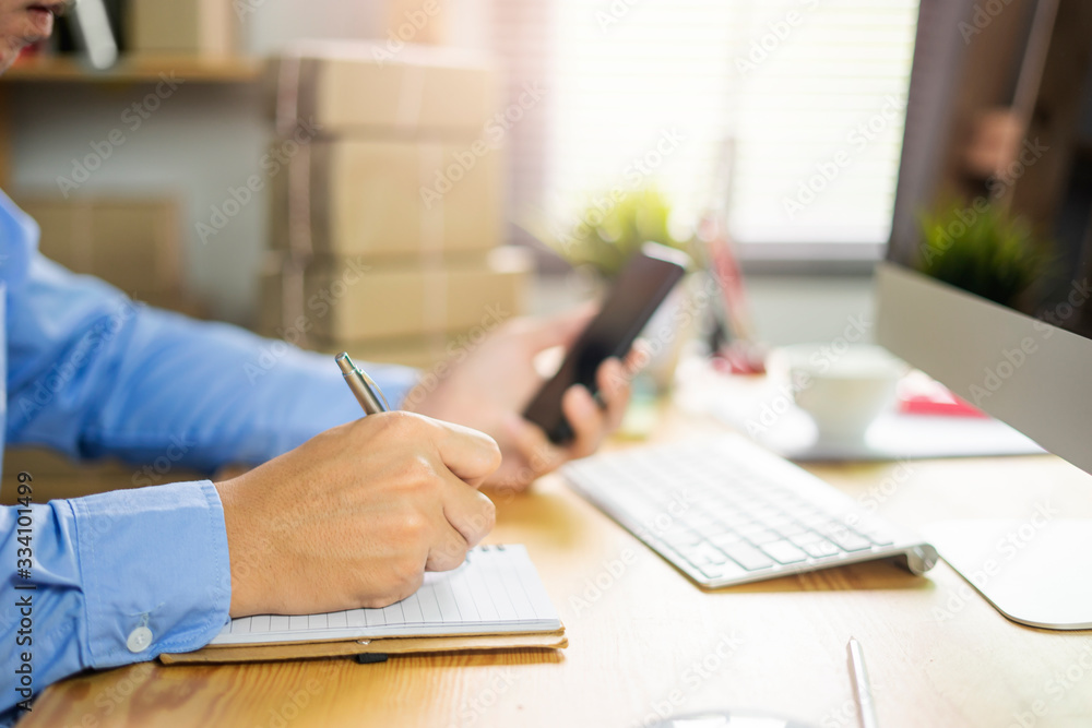 Close up of an asian man using notebook writing or drawing, planning or designing on work, while looking at phone, working in an office environment with clipboard of colour charts on computer desk