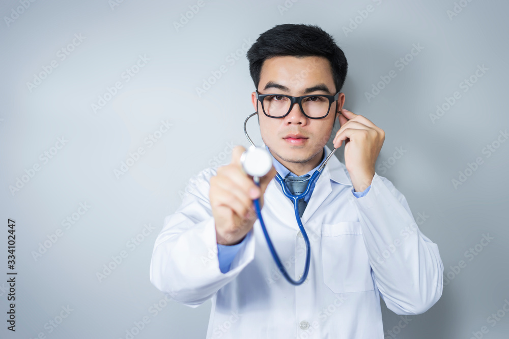 closeup portrait of a male asian doctor standing holding a stethoscope and listening to heart beat, thai ethnicity wearing glasses a white lab coat and blue shirt and tie, with bright grey background