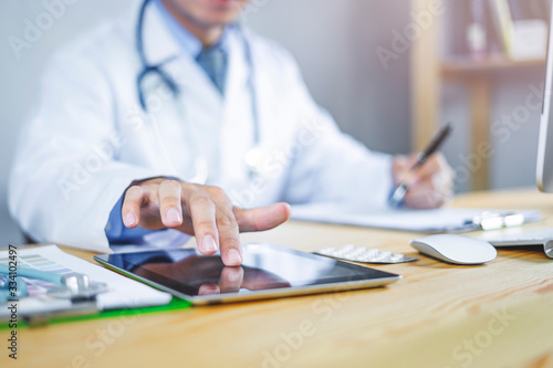 closeup concept of a male asian doctor touching a smart tablet and writing on a notebook sitting on his computer desk, wearing white lab coat with stethoscope around neck with sunshine in background