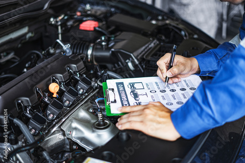 concept of a male asian engineer or a car mechanic looking inside car engine, holding a document on a clipboard diagnosing and checking parts to repair the car engine, wearing overall garnet and hat