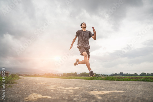 asian teen running along the road in sprinting action, exercising or practicing for a marathon race, wearing shirt, short and running shoes with mountain nature and cloudy sunset sky in the background