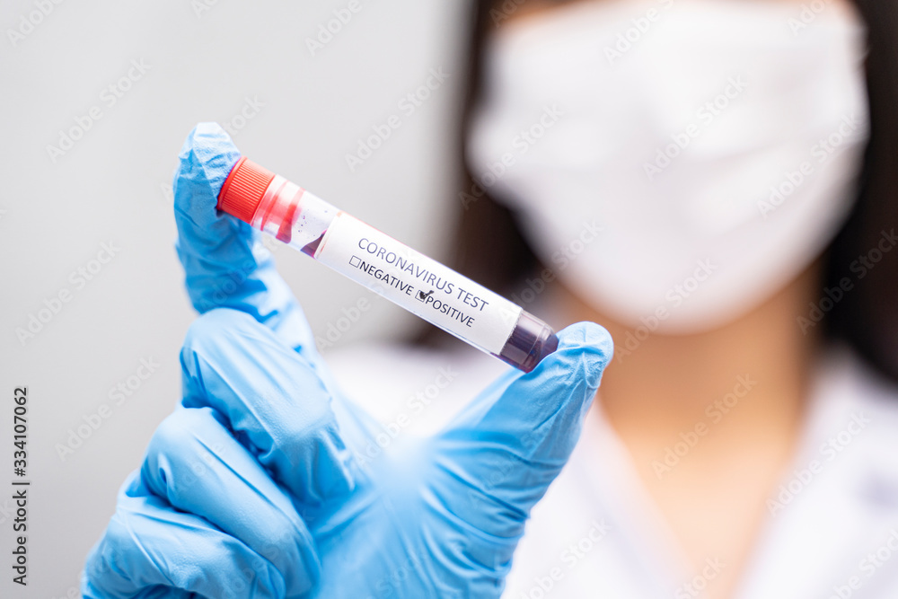 Close up of asian doctor holding a test tube blood sample of coronavirus wearing lab coat, white face mask, and blue nitrile gloves for protection against contagious virus infection, grey background Close up of asian doctor holding a test tube blood sample of coronavirus wearing lab coat, white face mask, and blue nitrile gloves for protection against contagious virus infection, grey background