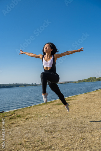 Fototapeta Naklejka Na Ścianę i Meble -  Smiling young female gymnast is jumping in split outdoors near the lake. Healthy lifestyle