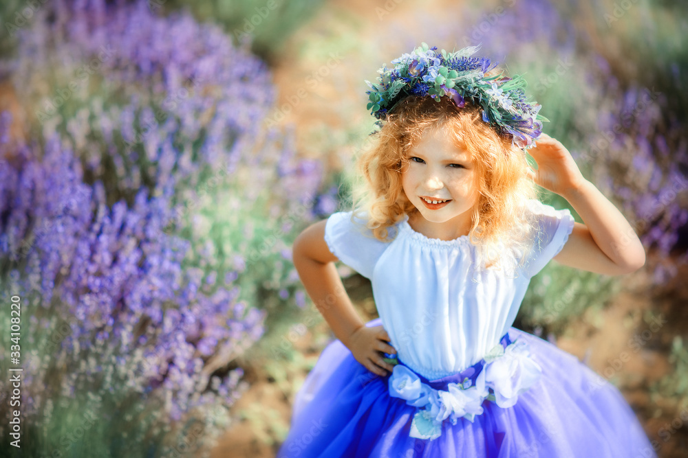 Fototapeta premium Portrait of happy cute little blonde curly girl wearing lavender dress and wreath in lavender field with violet flowers around in sunny summer day. Child in provencal landscape. Close to nature