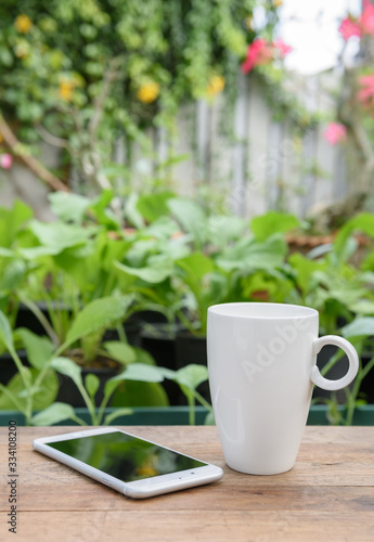 Mobile phone and white coffee cup on wood plank in vegetable farm and flowers garden background