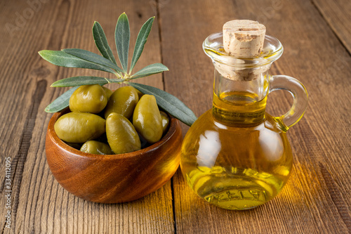 Green olives with olive branch in a small wooden bowl and a small bottle of olive oil on a rustic wooden background