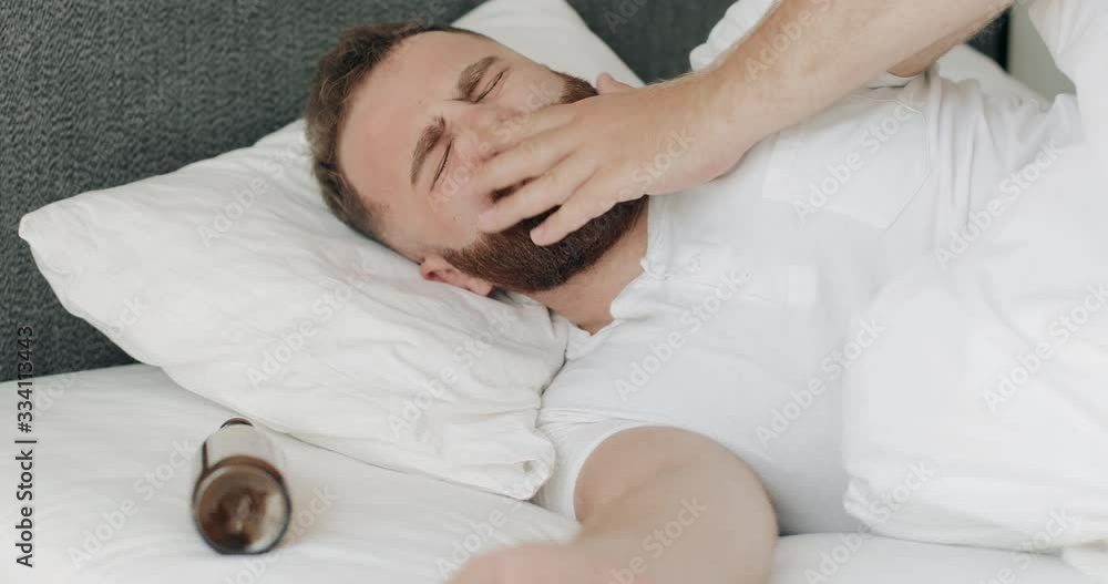 Young bearded man waking up while lying in bed with empty bottle near his head. Guy in 30s feeling bad and having headache after drinking alcohol and partying all night. Young bearded man waking up while lying in bed with empty bottle near his head. Guy in 30s feeling bad and having headache after drinking alcohol and partying all night.