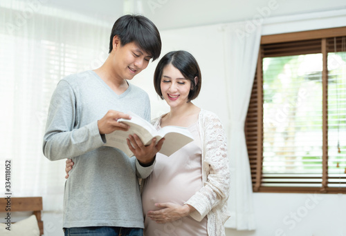 Young happy asian man and pretty pregnant woman reading book in home.Future father and pregnancy mother smile, laugh and enjoy together,family concept