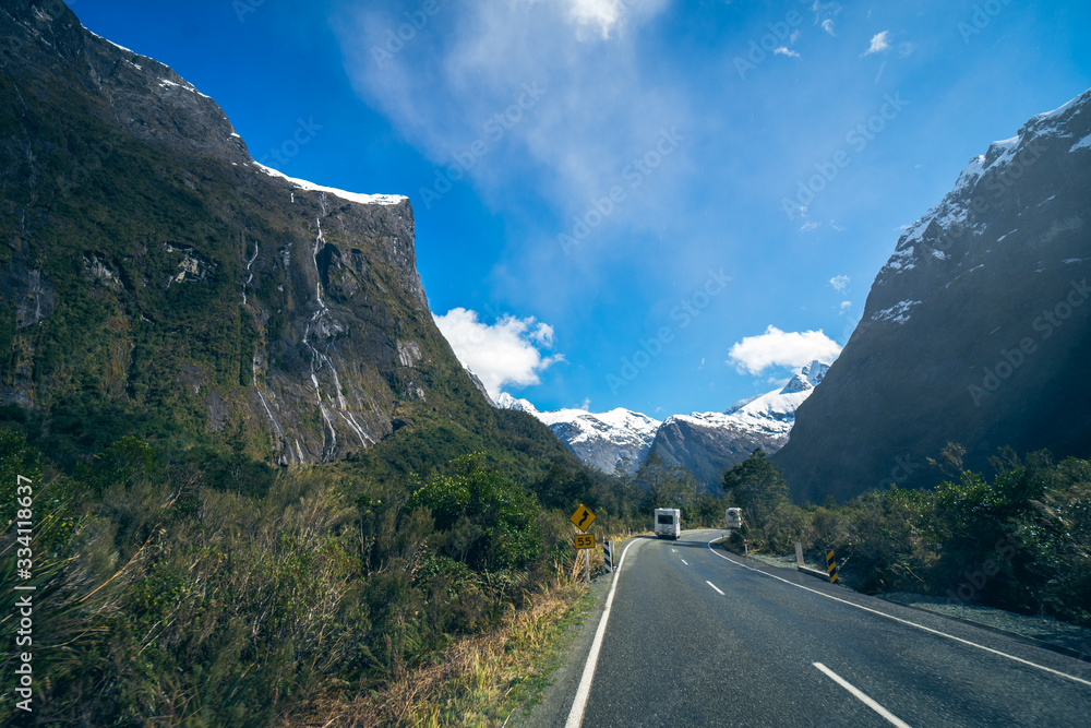Splendid image of the SH94 road towards Milford Sound surrounded by greenery with snow capped mountains in the background taken on a sunny winter day, New Zealand