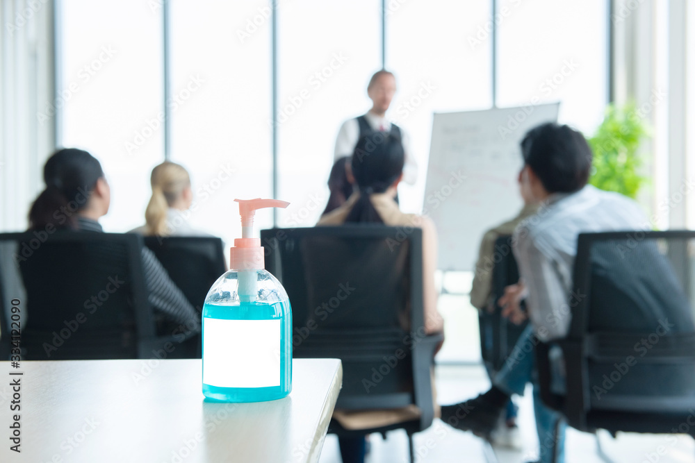 Alcohol gel bottle on table with blurred defocused group of meeting business person in background, health care personal hygiene in work place concept. Alcohol gel bottle on table with blurred defocused group of meeting business person in background, health care personal hygiene in work place concept.