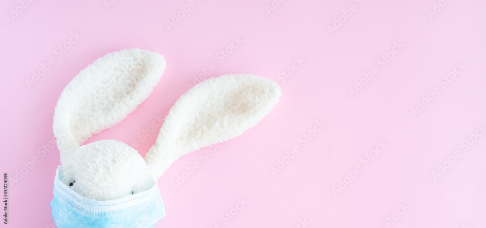 Easter quarantine concept, flat lay white bunny rabbit with face  protective mask on pink background. Copy space, top view.
