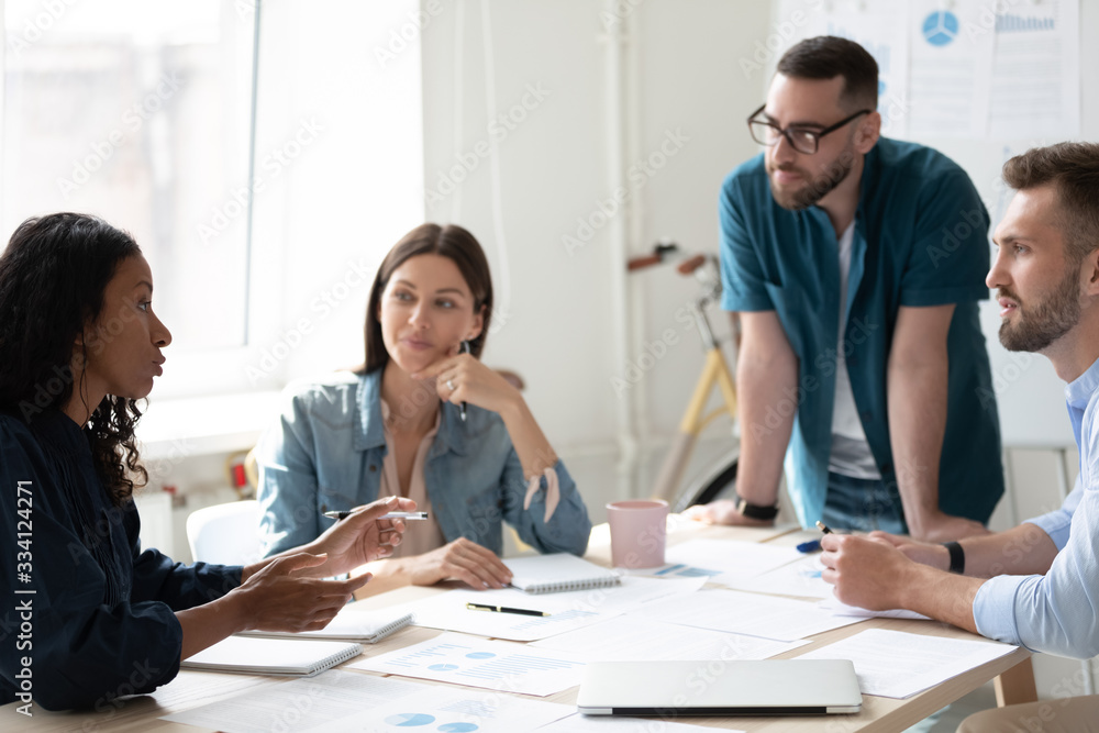 Fototapeta premium Motivated multiracial businesspeople sit at desk in boardroom discuss financial paperwork at briefing, focused diverse colleagues brainstorm analyze ideas at team meeting in office, teamwork concept