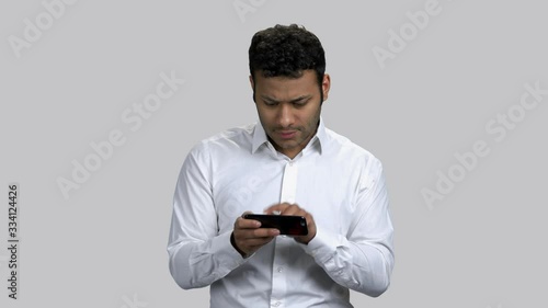 Infantile young dark-skinned man playing video game on his smartphone. Exctied hindu guy killing time, grey isolated background. photo