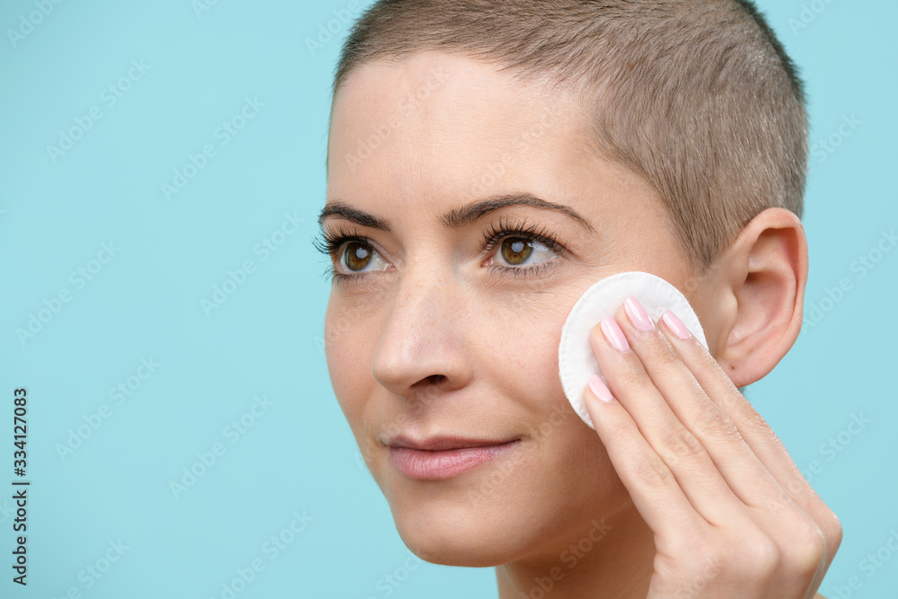 Smiling young woman removing make up using a cotton pad. Photo of an attractive caucasian woman with healthy skin and short haircut isolated on pastel blue background. Beauty and Skincare concept.