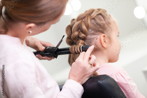 The work of the stylist on hairstyles in a bright salon, the girl makes curls and braids braids of a small beautiful model,