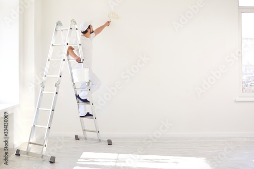 A male painter in a white uniform with a roller works in his hand in a white room