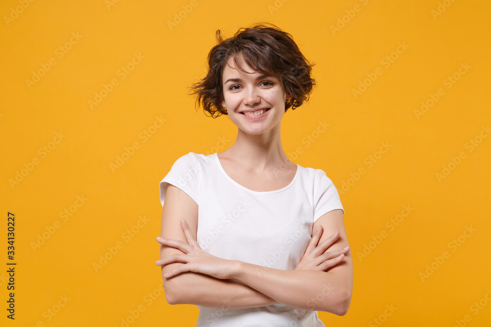 Obraz premium Smiling young brunette woman girl in white t-shirt posing isolated on yellow orange background studio portrait. People sincere emotions lifestyle concept. Mock up copy space. Holding hands crossed.