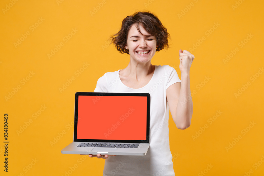 Smiling young brunette woman in white t-shirt posing isolated on yellow background. People lifestyle concept. Mock up copy space. Hold laptop pc computer with blank empty screen doing winner gesture. Smiling young brunette woman in white t-shirt posing isolated on yellow background. People lifestyle concept. Mock up copy space. Hold laptop pc computer with blank empty screen doing winner gesture.