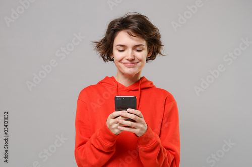 Smiling young brunette woman girl in casual red hoodie posing isolated on grey wall background studio portrait. People lifestyle concept. Mock up copy space. Using mobile phone, typing sms message.